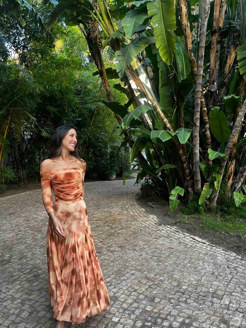 Vestido largo de The IQ Collection con escote bardot, manga larga y estampado difuminado naranja, con gargantilla dorada, en un jardín para invitada de boda de día