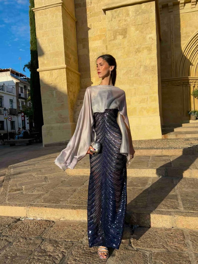 Vestido largo azul noche de lentejuelas con capa de satén gris plata, delante de una iglesia de piedra