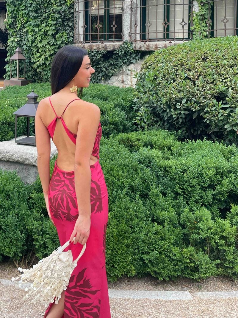 Vestido largo fucsia de Lady Pipa con hojas estampadas en granate, tirantes cruzados y la espalda al aire, con bolso pequeño blanco, de espaldas en un jardín, para una graduación