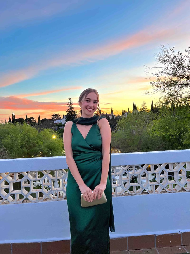 Vestido largo de satén verde botella de The Are con capa a juego al cuello y clutch dorado, al atardecer en una terraza