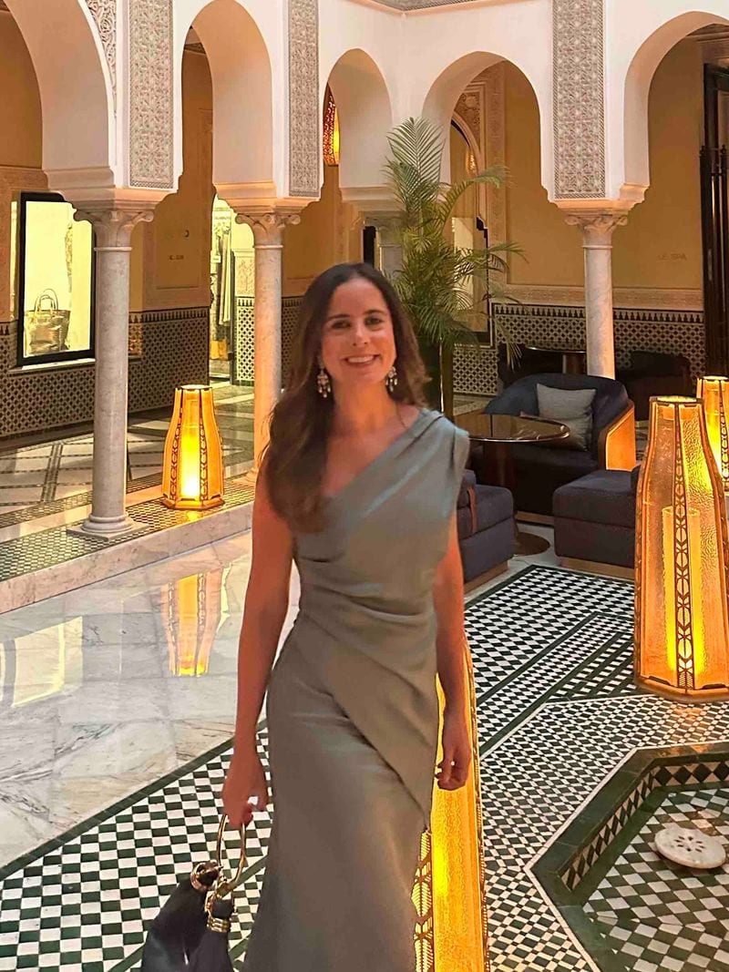 Vestido gris verdoso de un hombro con la tela recogida en la cintura, con pendientes largos dorados y bolso negro, en un patio con arcos y faroles al anochecer, para invitada de boda de otoño