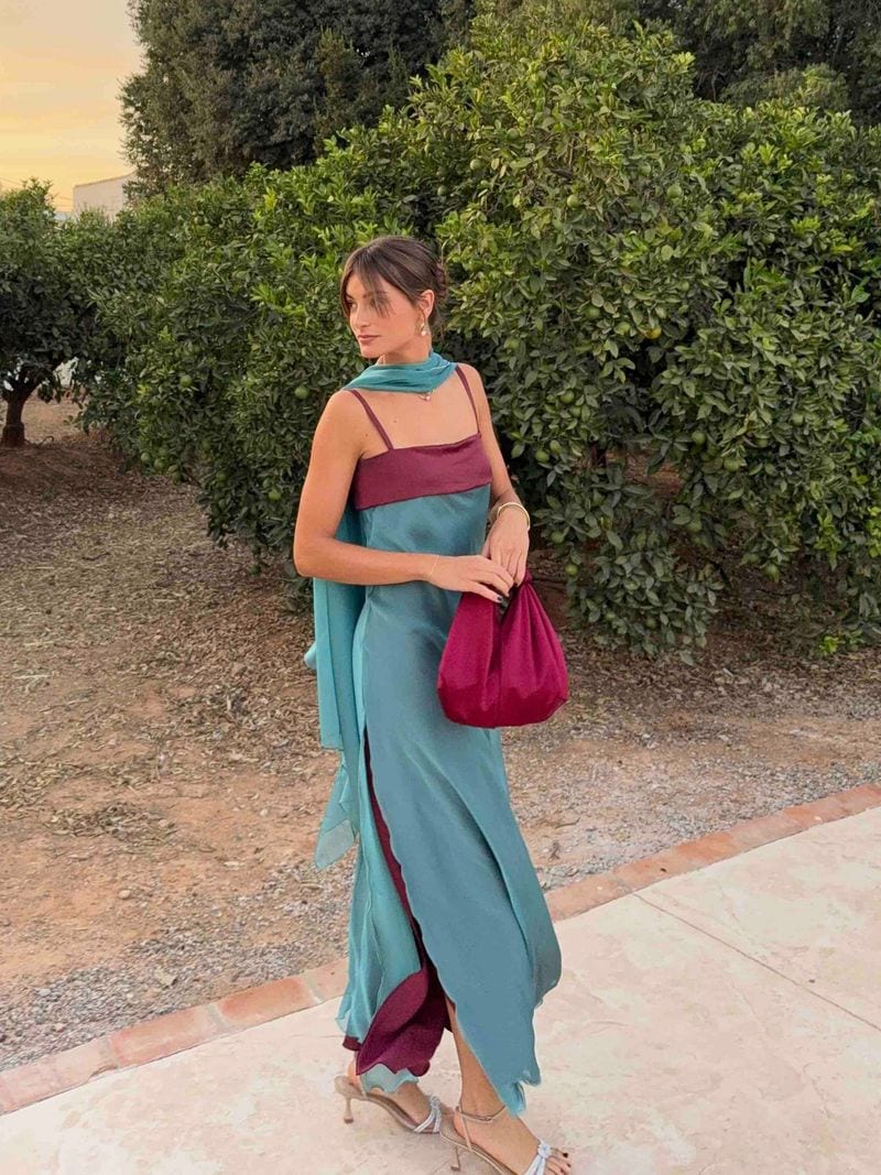 Vestido lencero turquesa con banda granate en el escote y capa turquesa anudada al cuello, con bolso de satén granate y sandalias plateadas, entre naranjos al atardecer, para invitada de boda en una finca