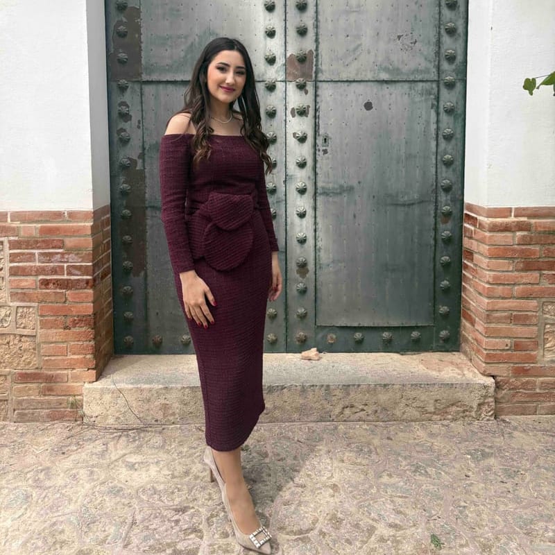 Invitada con vestido midi burdeos de tweed de Coosy, de hombros al aire, manga larga y nudo de flor en la cintura, delante de un portón antiguo